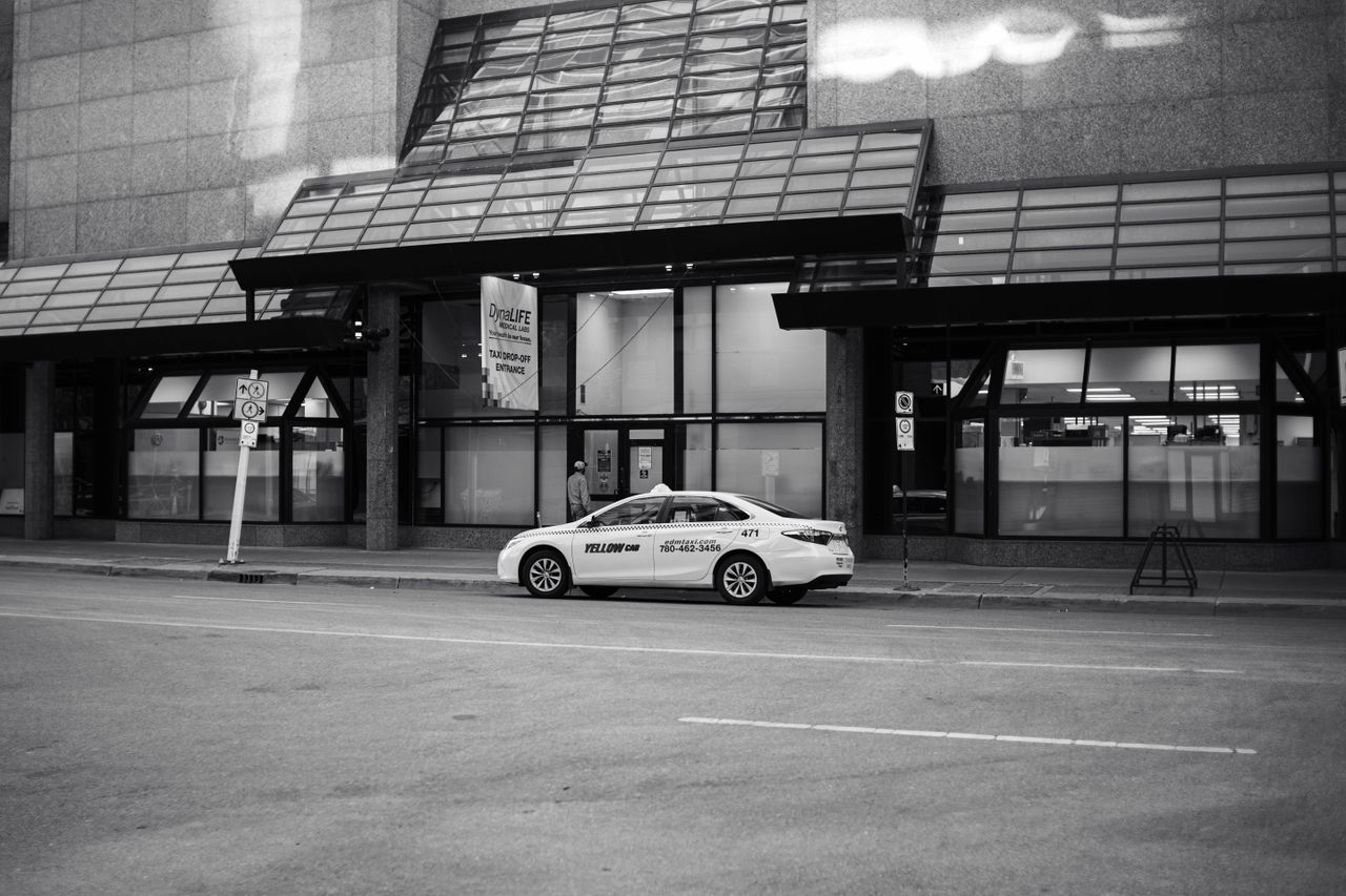 Black and white urban street scene featuring a parked taxi by modern glass buildings.
